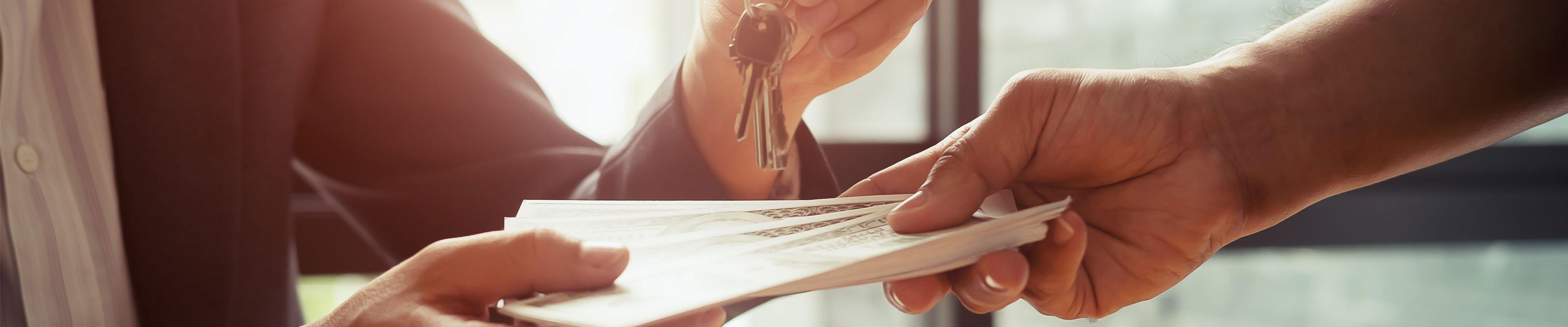 A white woman in a nice suit takes cash from a white man in exchange for a house key. The image is cropped to only their hands.