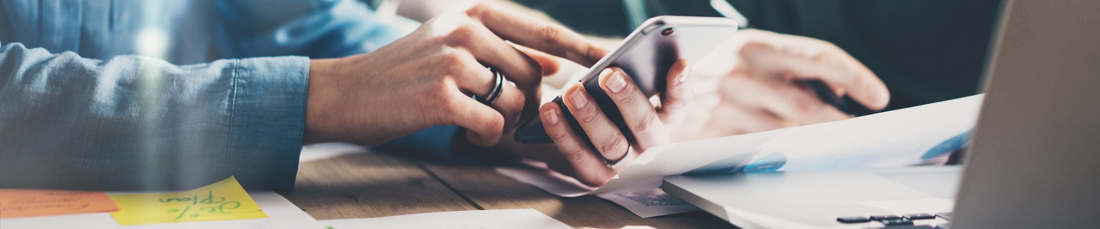 A white man wearing a black ring on his right ring finger is typing on a silver smartphone while reviewing pie charts.